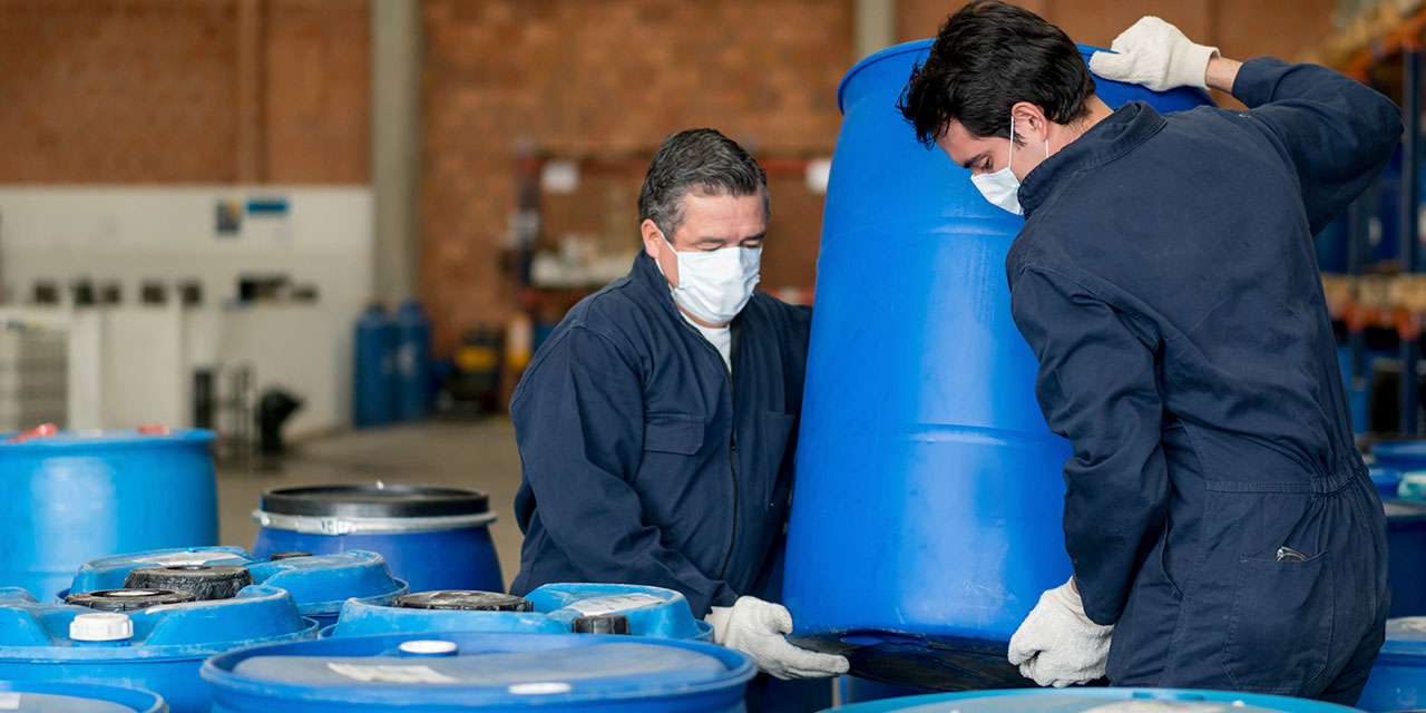 Industrial workers wearing protective masks and gloves lifting large blue chemical drums in a warehouse facility.