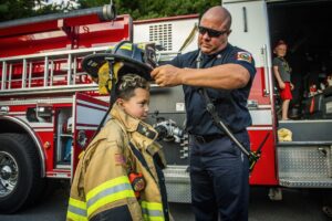 A child trying on adult firefighter gear, aided by a fireman
