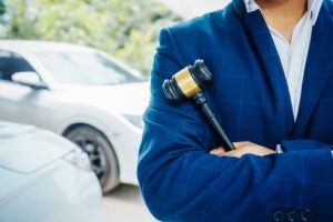 Hombre de traje sosteniendo un mazo, con coches al fondo.