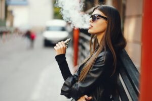 A fashionable woman strolls through the urban landscape, puffing on an e-cigarette with effortless grace.