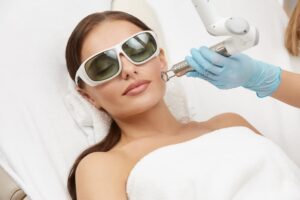 In a spa salon, a woman gets laser treatment on her cheek while wearing glasses. A beautician administers facial procedures.