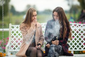 Beautiful teenage girls in casual attire sit on a vintage bench on the street, smoking and vaping electronic cigarettes in the evening.