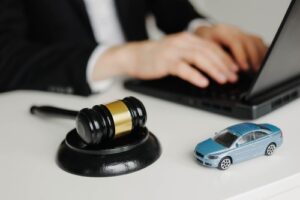 Judge’s gavel and toy car on a desk with a person typing on a laptop.