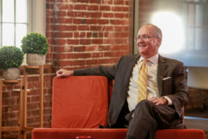 A professional man wearing a suit and tie sits comfortably on a red sofa, smiling, in a cozy office setting with a rustic brick wall background and natural light streaming in from a window.