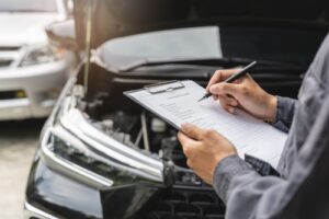 Mechanic writing on a checklist clipboard in front of a car with the hood open.