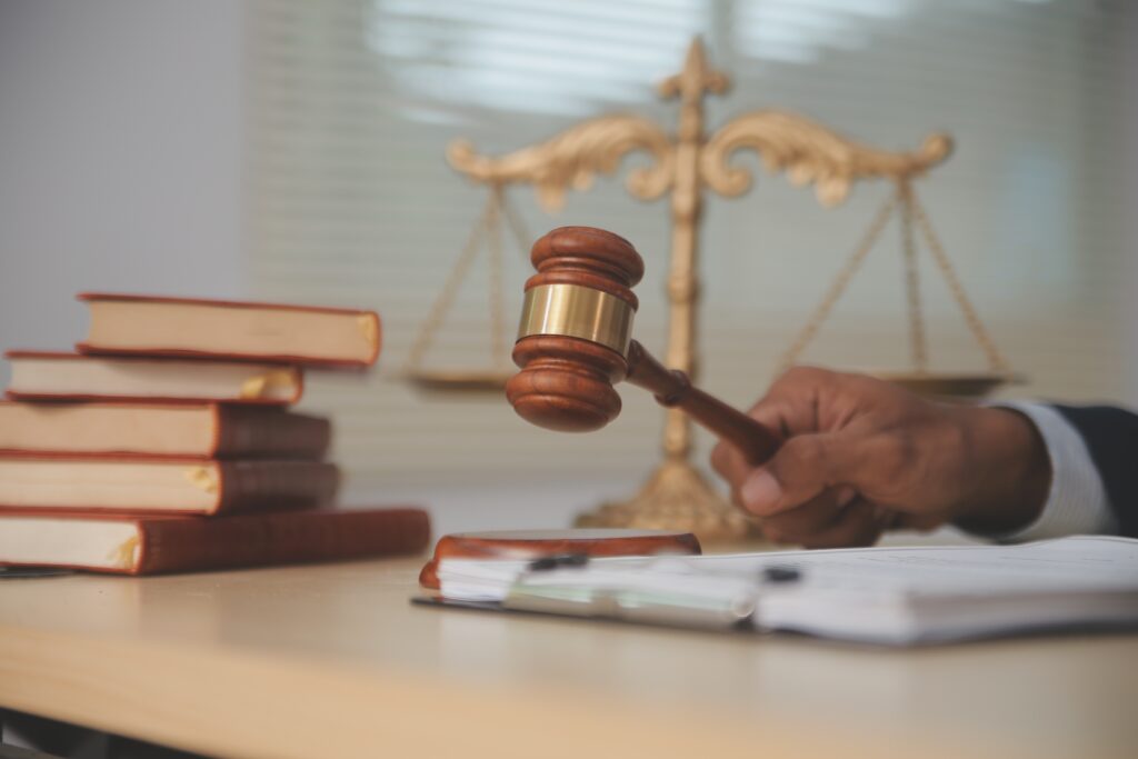 Judge’s gavel and legal books on desk with scales of justice in background during court proceedings. Judge’s gavel and legal books on desk with scales of justice in background during court proceedings.