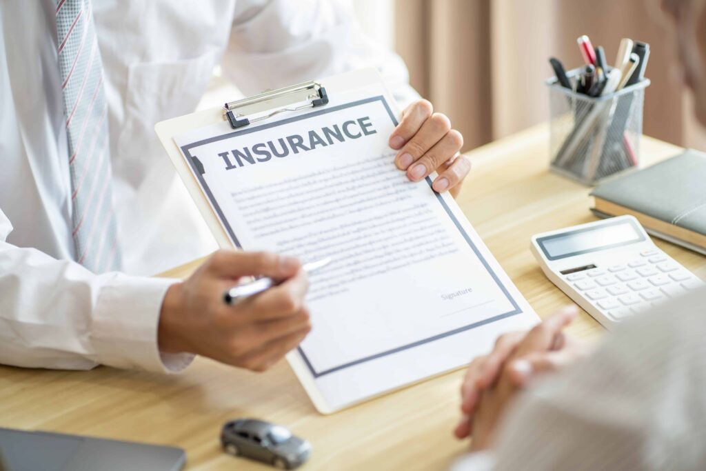 Insurance agent explaining policy details to a client with a contract on a clipboard at a desk.