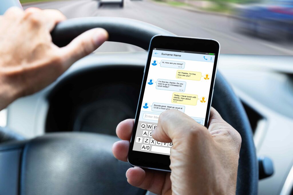 Driver using a smartphone to send text messages while driving, holding a phone over a steering wheel on a busy road. Driver using a smartphone to send text messages while driving, holding a phone over a steering wheel on a busy road.