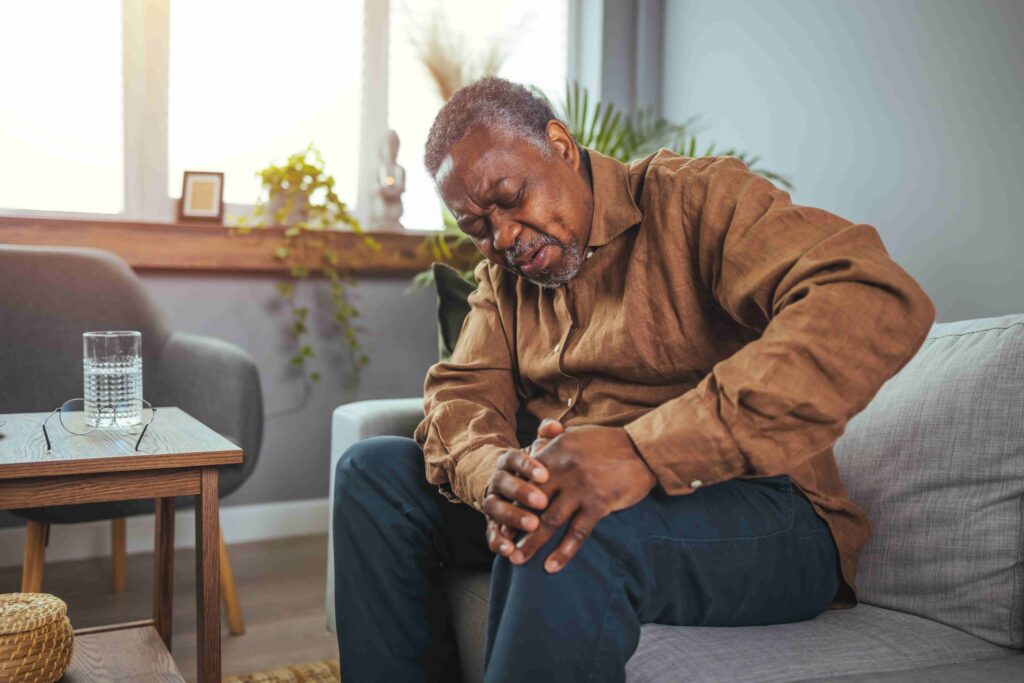 Senior man sitting on couch holding painful knee at home, illustrating knee injury and chronic joint pain for personal injury claim.