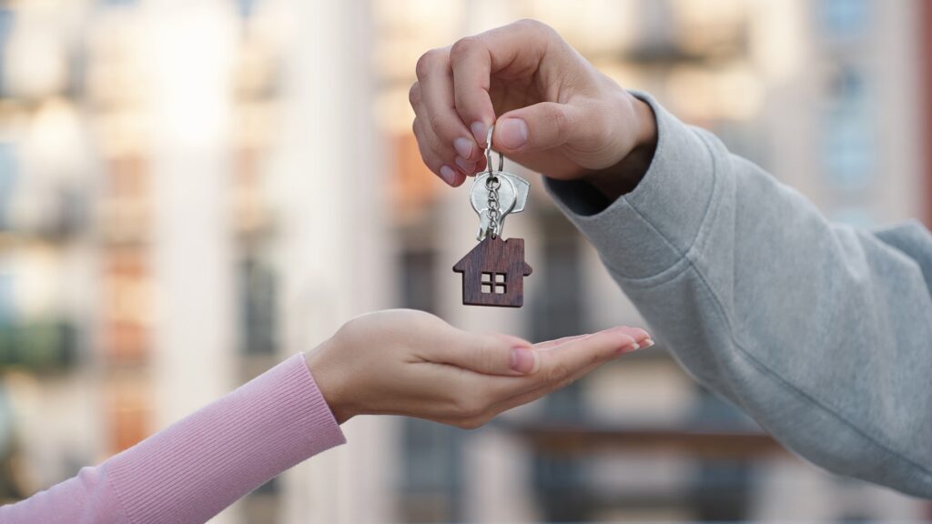 Person handing house keys with a home-shaped keychain to another person, symbolizing property transfer, home purchase, or real estate closing. Person handing house keys with a home-shaped keychain to another person, symbolizing property transfer, home purchase, or real estate closing.