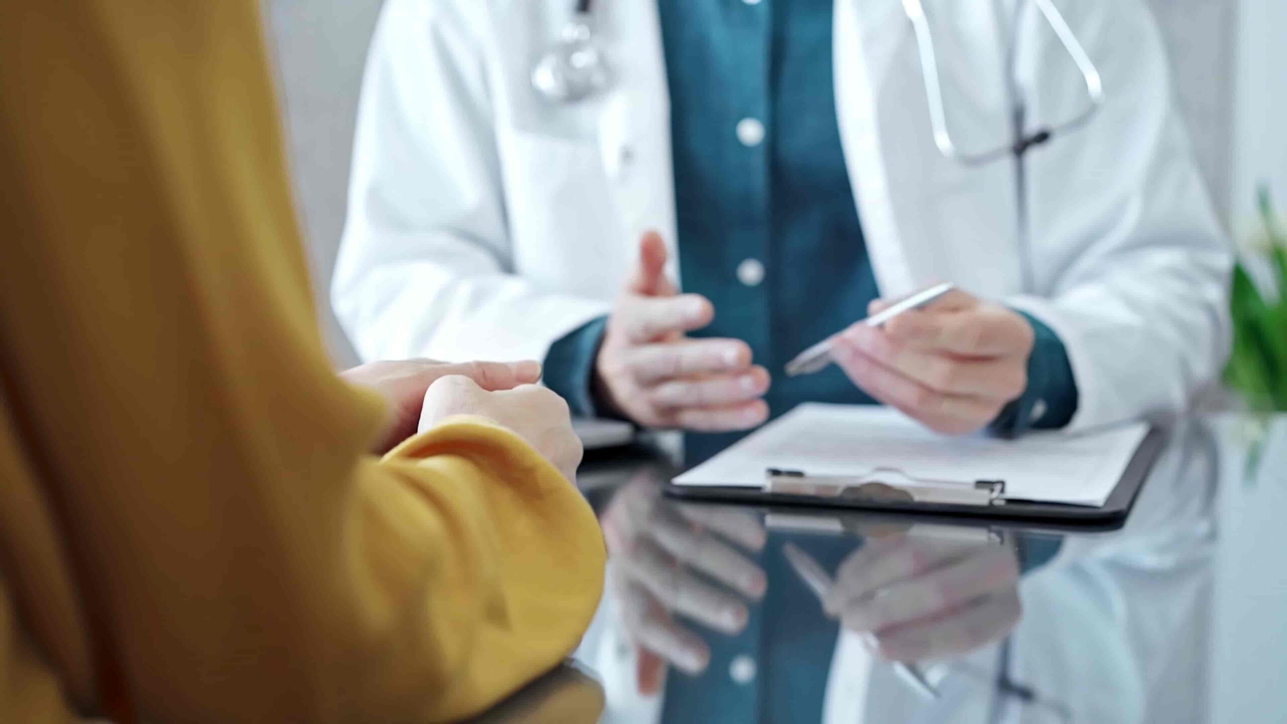 Médico consultando con un paciente en un escritorio, sosteniendo un portapapeles, discutiendo su condición médica, opciones de tratamiento y consejos de salud en un entorno clínico.